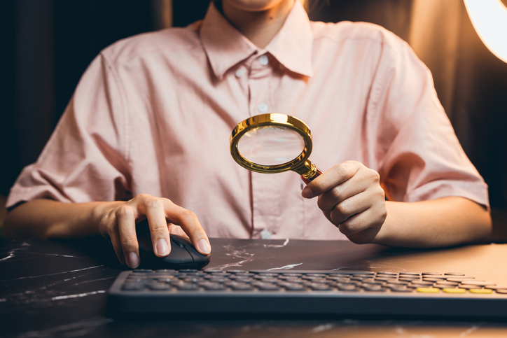 Person Holding Magnifying Glass While Using Computer Mouse in Office Environment for Investigation or Analysis alternate text for this image