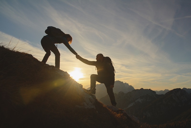 A mid-adult man and woman clad in hiking attire exchange a joyful high five on a mountaintop, silhouetted against a serene sunset sky.