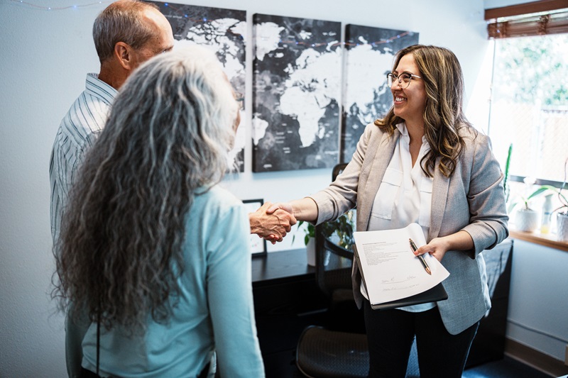 A smiling Latina financial advisor stands in her office and shakes hands with a senior couple following a retirement planning consultation.