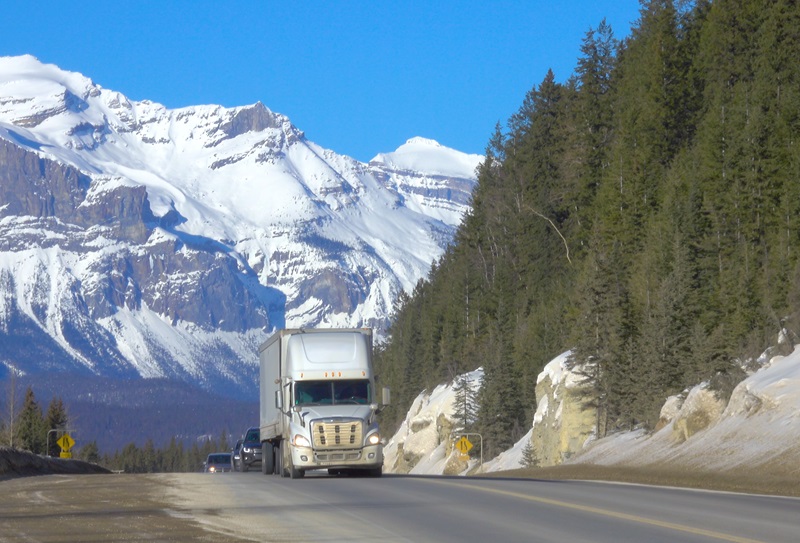 Big 18 wheeler and cars drive down the famous Icefields Parkway on a sunny spring day. Traffic moves along an interstate highway running under the snowy mountains. Road trip across Rocky Mountains.